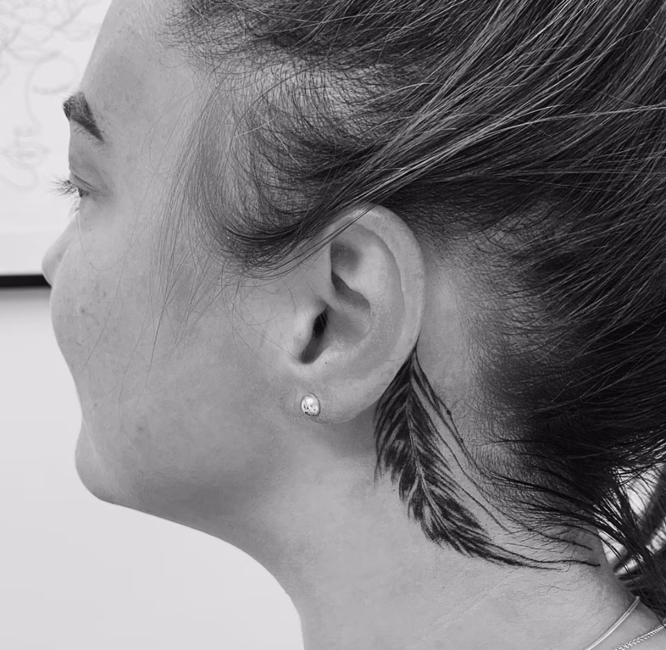 Black fine-line feather tattoo behind a client’s ear by Daria at Calypso Beauty