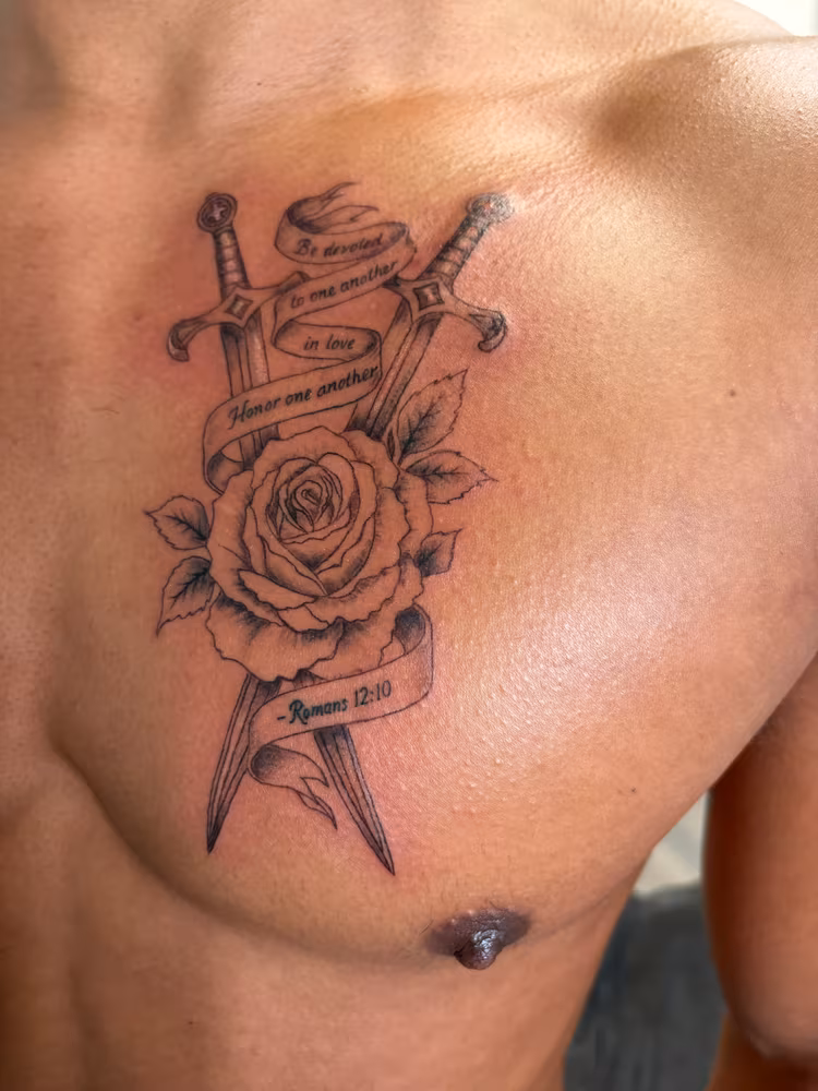 Chest tattoo of a shaded rose and crossed swords with a scripture banner reading Romans 12:10